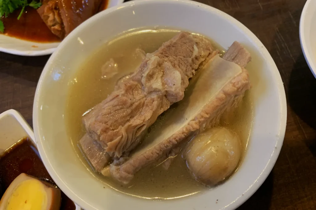 A white bowl of clear soup containing tender pork ribs, a large bone, and a whole onion, arranged on a wooden table for a comforting meal.