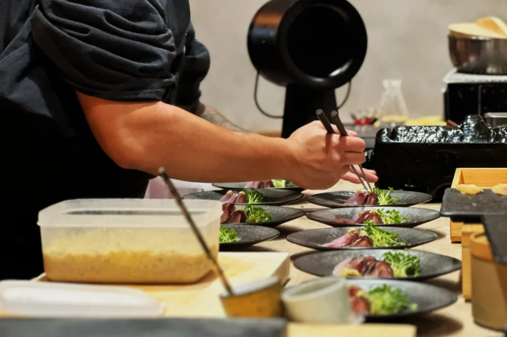 Chef Masa artfully arranges food on multiple black plates with precision, using tweezers. The scene is organized, conveying a sense of focus and expertise.