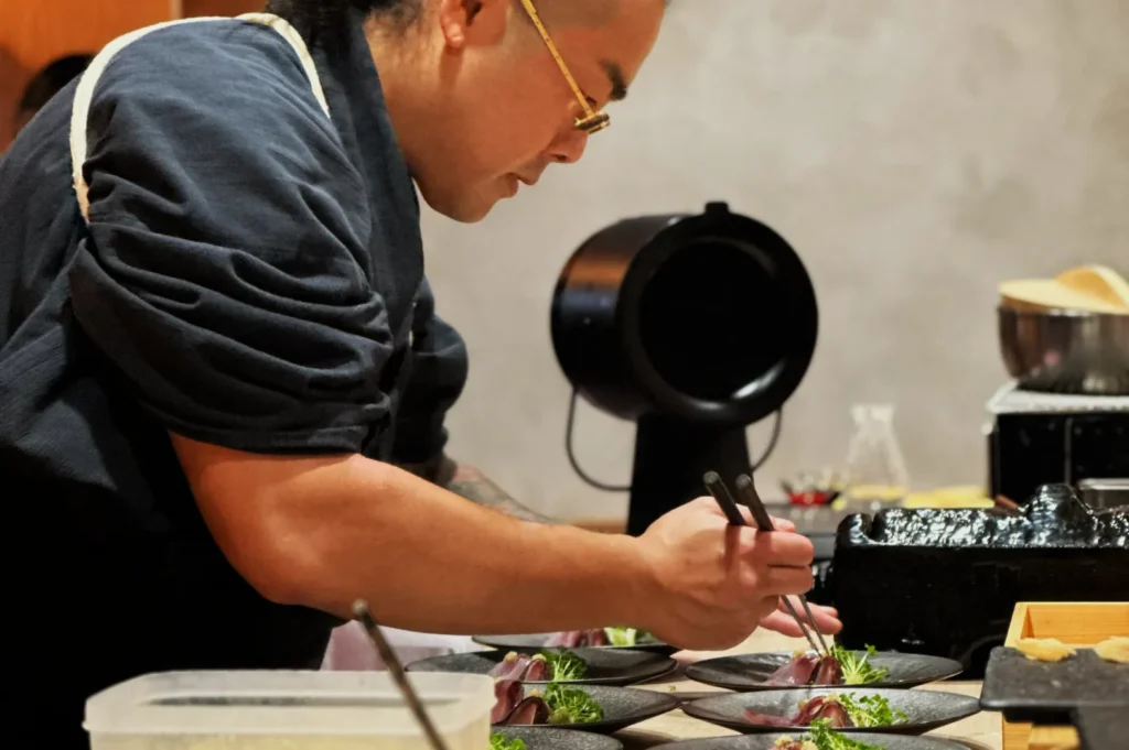 A chef in glasses and a dark apron attentively prepares dishes with tweezers, focusing on presentation. The atmosphere is professional and calm.