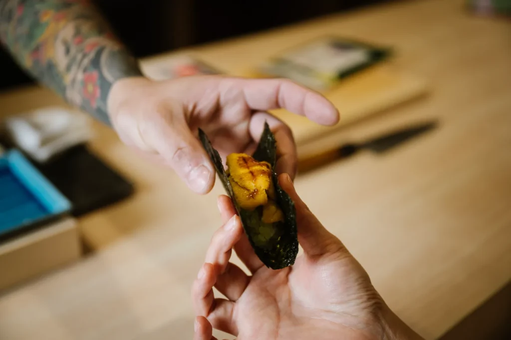 Two hands exchanging a piece of sushi wrapped in seaweed, with a focus on the uni topping. The background shows a wooden table and a blurred kitchen scene.