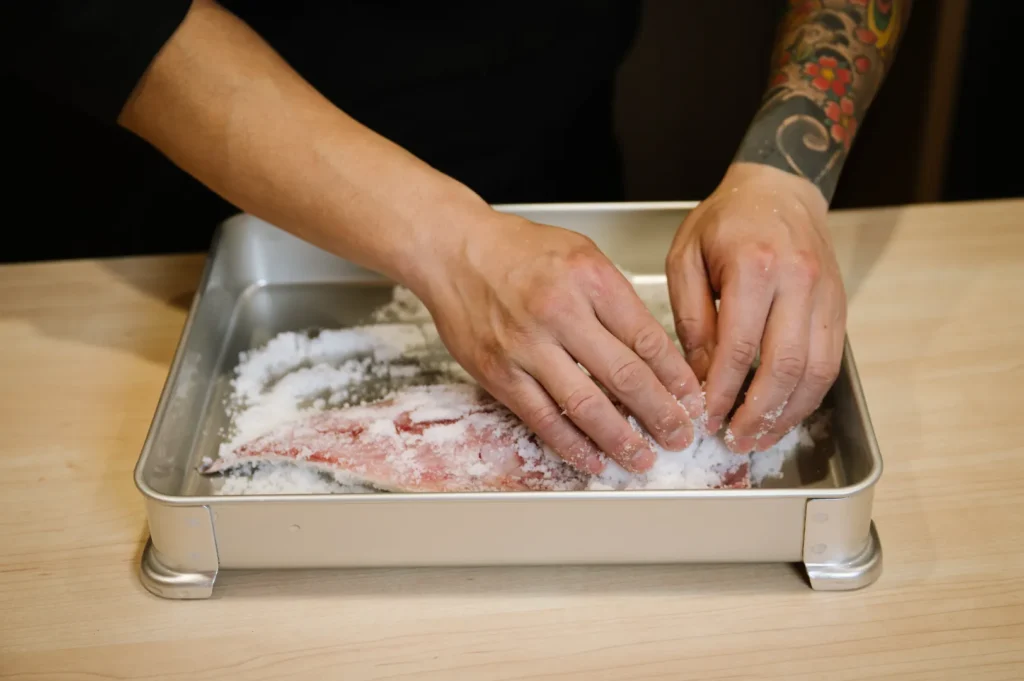 Chef Masa with tattooed arms is salting a fish fillet in a shiny metal tray on a wooden table, highlighting skillful preparation in cooking.