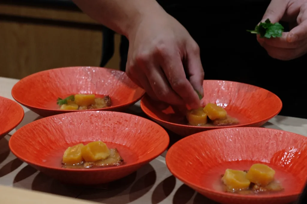 Chef arranging herbs on vibrant orange plates with sliced fruit and sauce, showcasing meticulous presentation and culinary artistry.