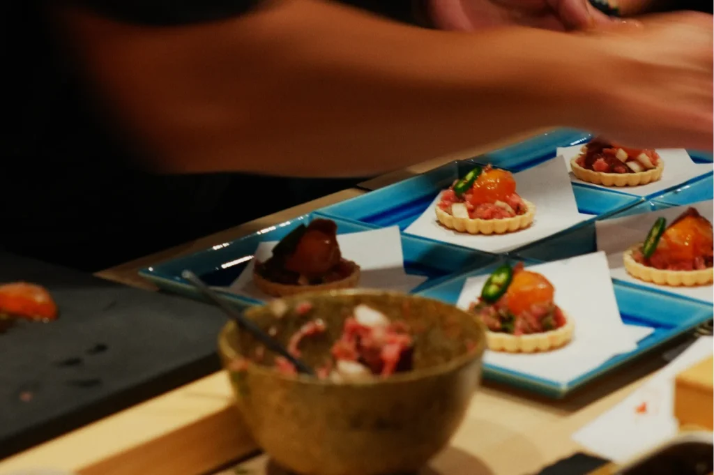 Close-up of a chef's hands arranging tartlets with toppings on a blue plate. A bowl of mixed ingredients sits nearby, suggesting preparation and care.