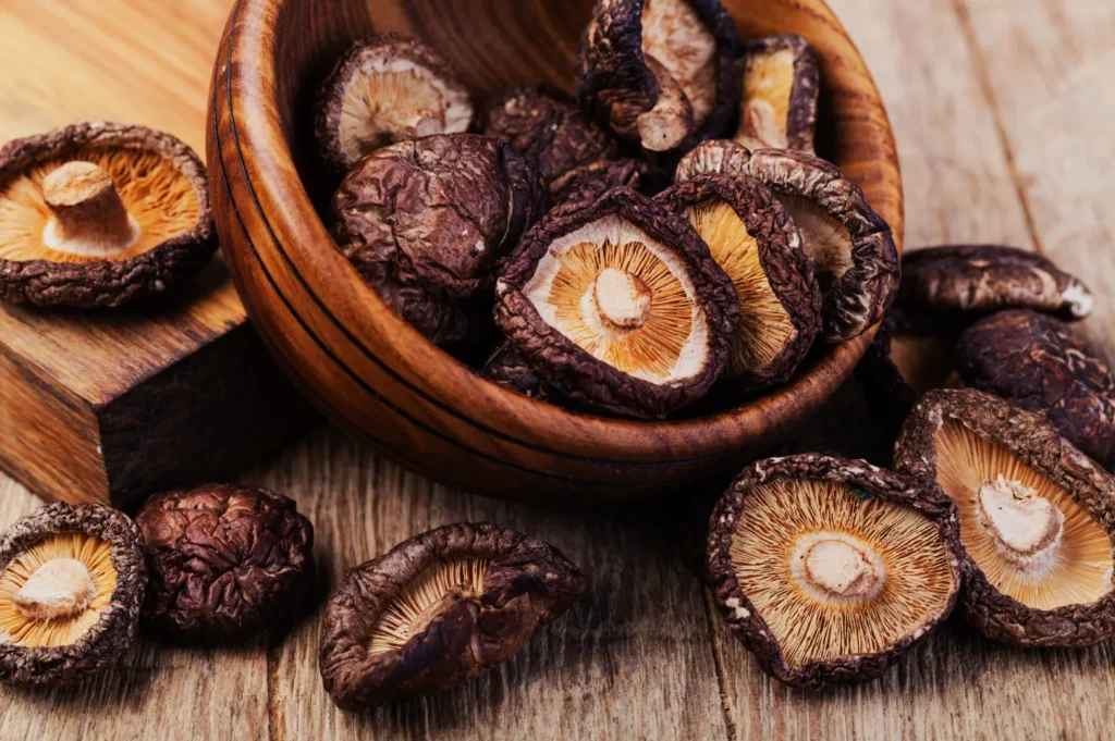 A wooden bowl overflows with dried shiitake mushrooms on a rustic wooden table. Some mushrooms spill out, highlighting their textured caps and stems.