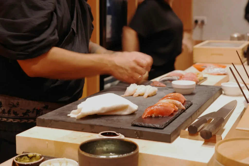 Chef preparing sushi on a wooden counter. Various types of sushi are arranged on a black slate, with bowls and knives nearby, creating a focused and skilled ambiance.