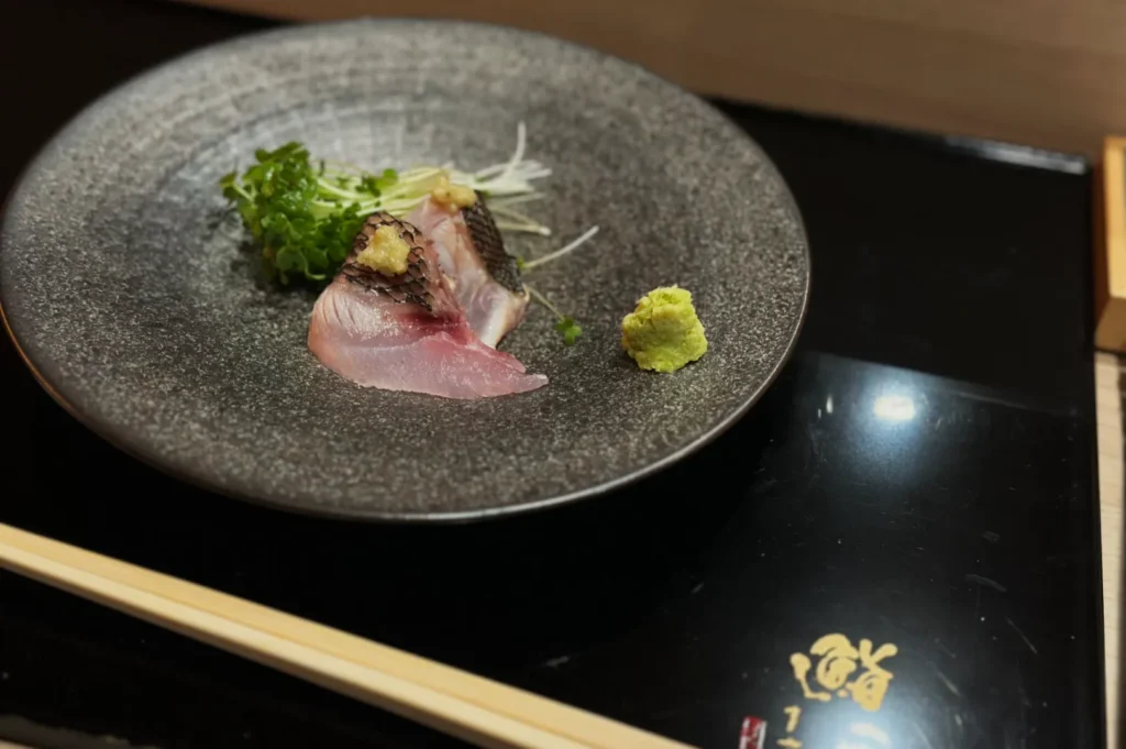 A grey plate with two slices of sashimi, garnished with greens and ginger, and a wasabi mound. Chopsticks rest nearby on a dark surface.