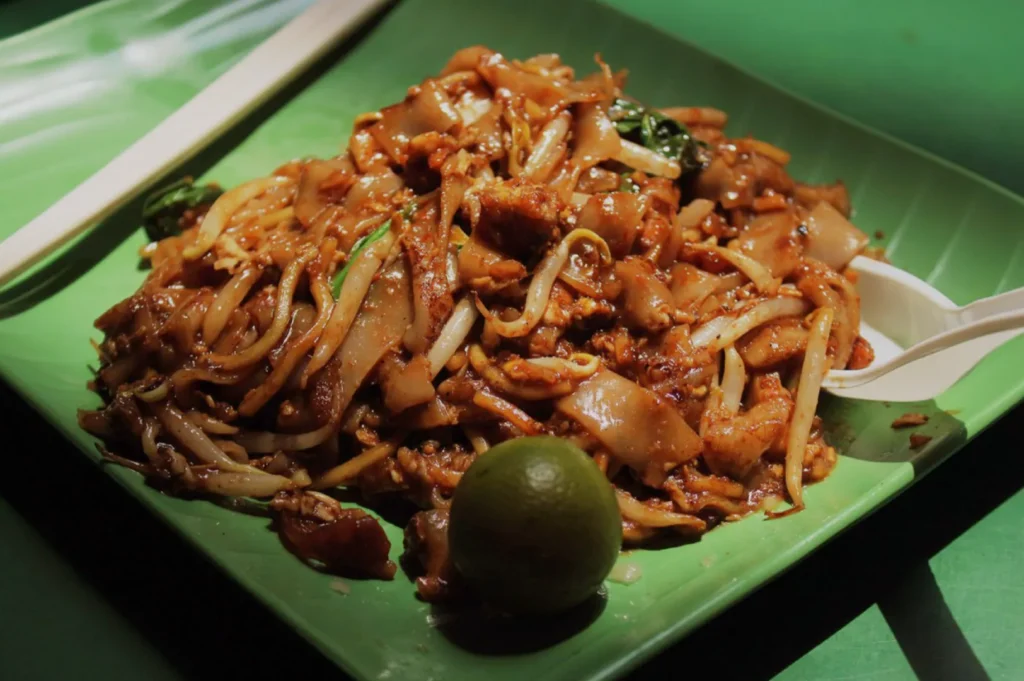 A vibrant plate of stir-fried noodles with chicken and vegetables on a green dish. A lime wedge and plastic utensils are beside the dish.