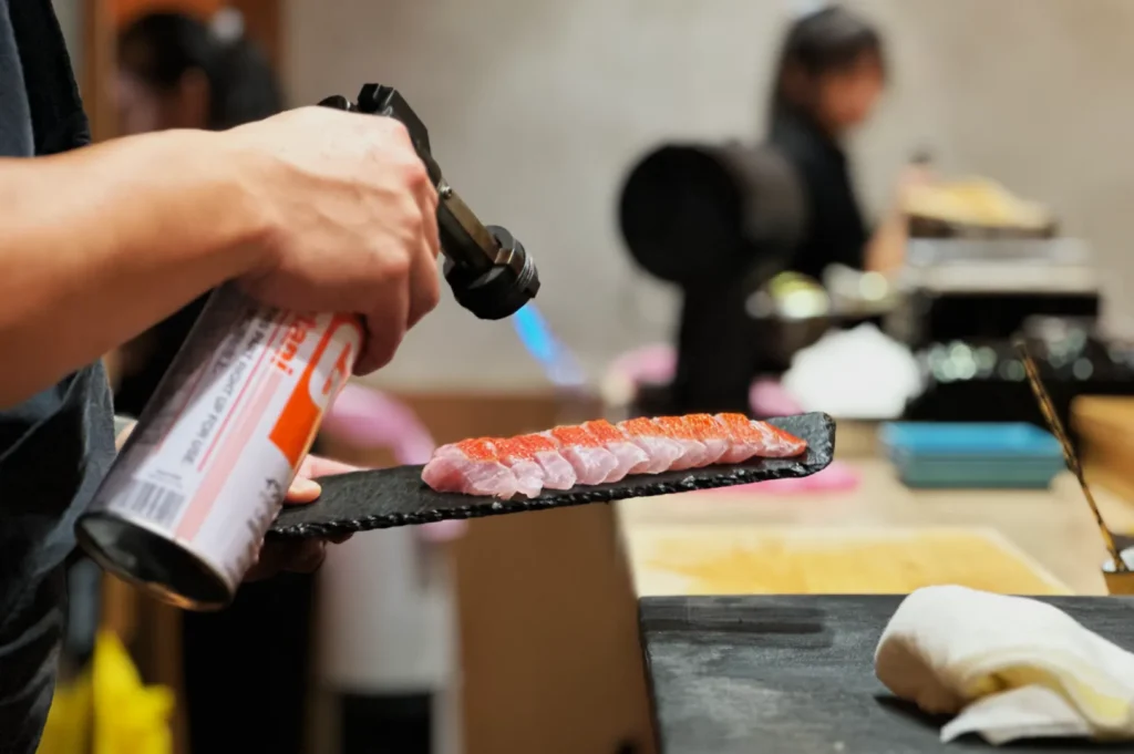 Chef Masa uses a blowtorch to sear sushi on a black tray. The background shows a blurred kitchen setting, creating a focused and professional atmosphere.