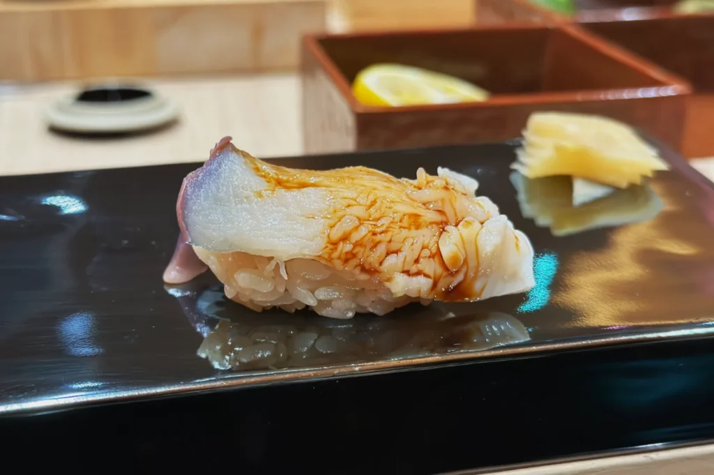 Sushi piece with fish and sauce on black plate with sliced ginger. Wooden boxes and lemon visible in background. Elegant and appetizing presentation.