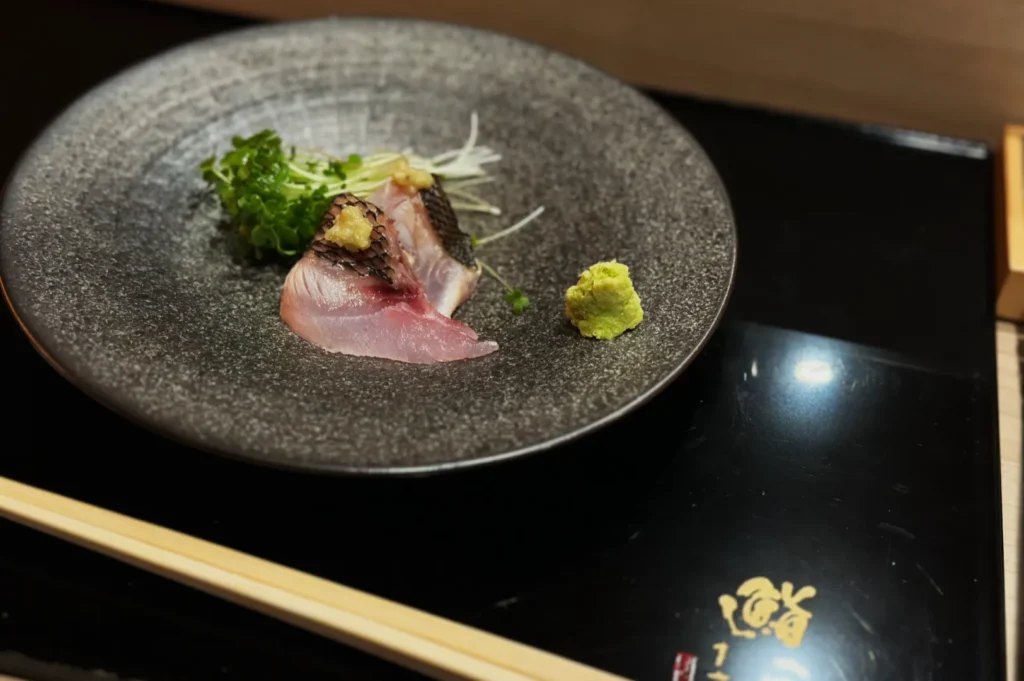 A dish of sashimi garnished with greens and ginger on a textured gray plate. A small mound of wasabi sits beside it, with chopsticks nearby.