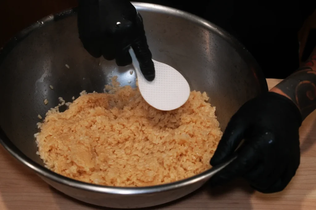 Person wearing black gloves mixing rice with a white spatula in a large metal bowl, creating a focused and methodical cooking scene.