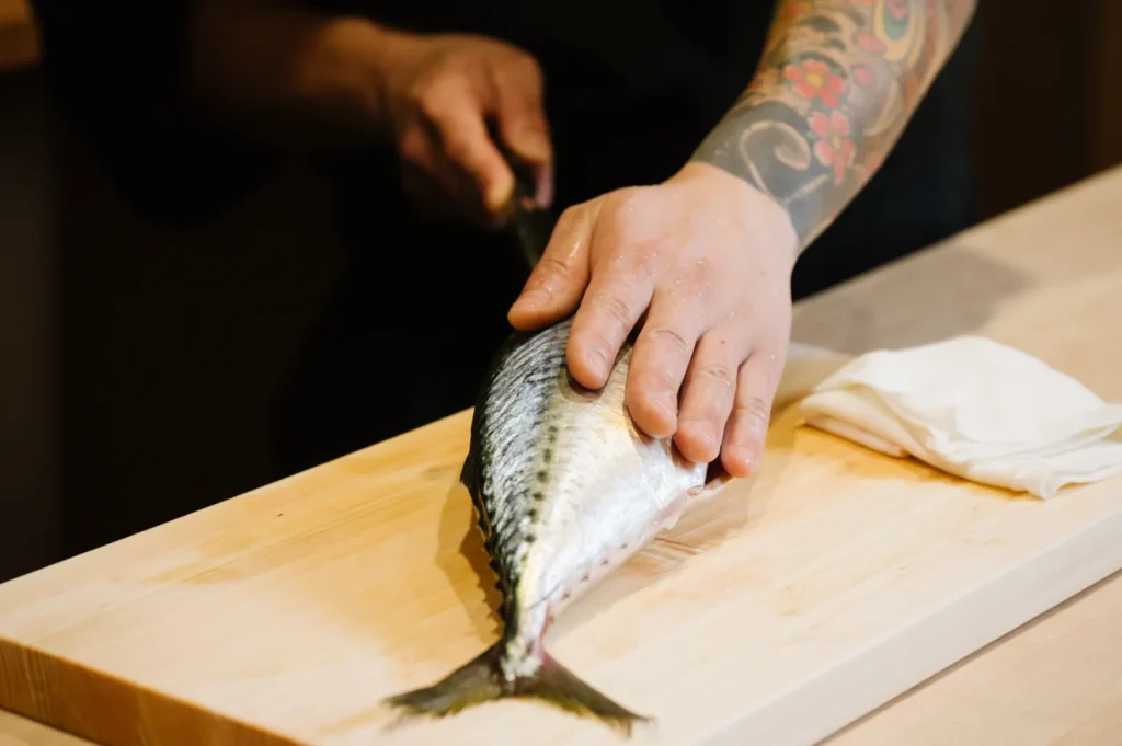 Tattooed chef Masa's hands filleting a mackerel on a wooden cutting board, showcasing culinary skill. A folded cloth is placed beside the fish, adding to the kitchen setting.