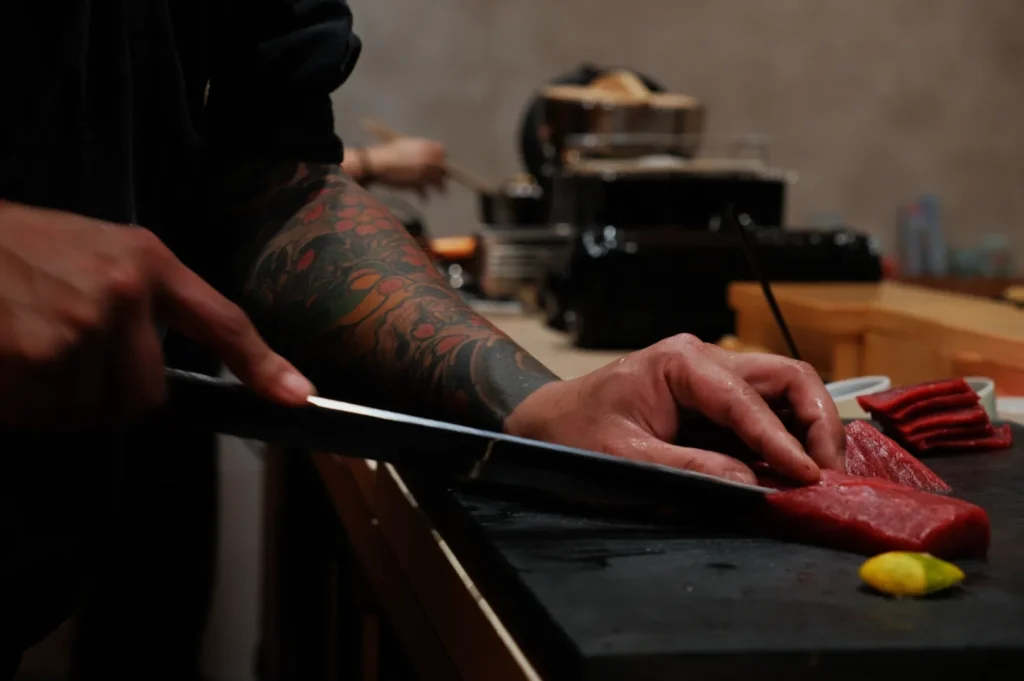 Close-up of a tattooed chef slicing raw tuna with a sharp knife. A lime wedge and more tuna pieces are nearby. The scene feels focused and precise.