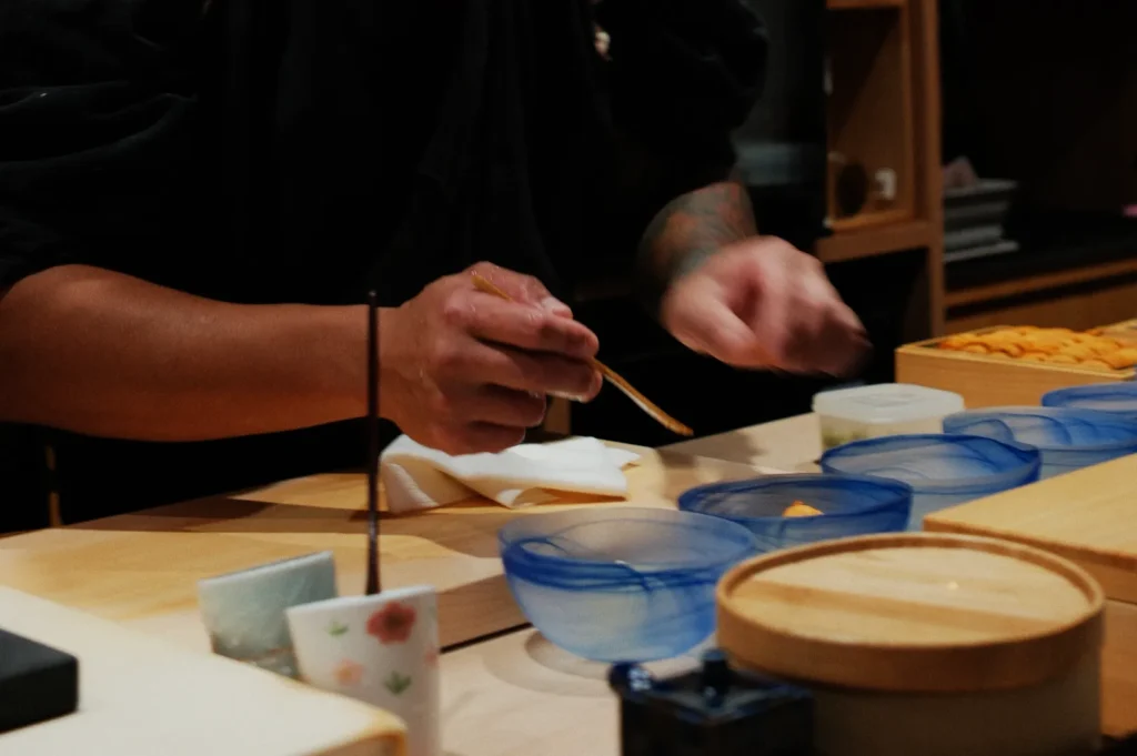 Chef skillfully prepares food using chopsticks at a kitchen counter, surrounded by blue bowls and ingredients, creating an atmosphere of focus and precision.