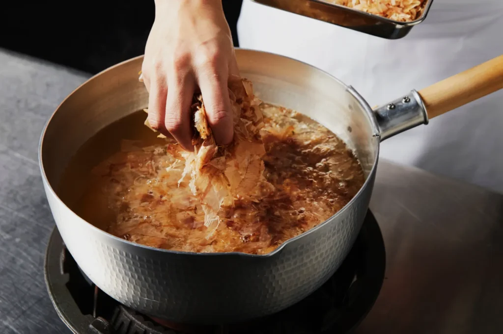 A hand adds katsuobushi (bonito flakes) to a simmering pot of broth on a stove, suggesting a calm cooking process in a kitchen setting.