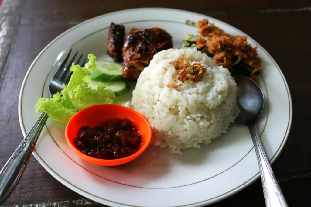 A plate of Indonesian food featuring white rice topped with fried shallots, grilled chicken, green vegetables, and a small orange bowl of sambal.