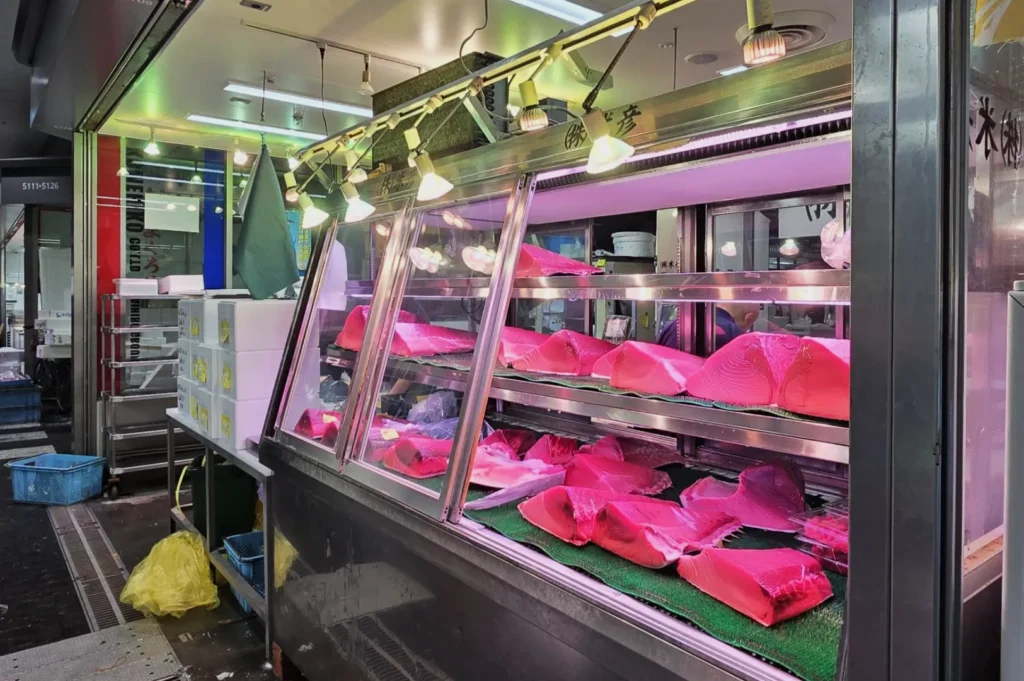 Refrigerated seafood display with vibrant pink tuna cuts on green mats, under bright lights in a market setting. Cool, fresh ambiance with metallic surfaces.