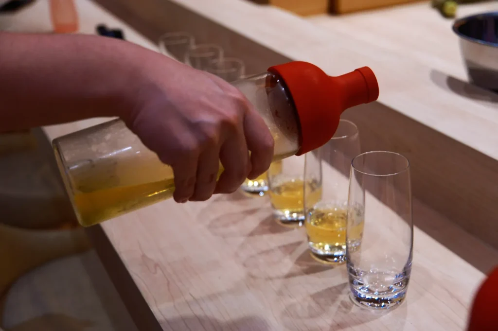 A hand pours a yellow liquid from a bottle with a red spout into tall glasses lined up on a wooden counter, suggesting a preparation for serving drinks.