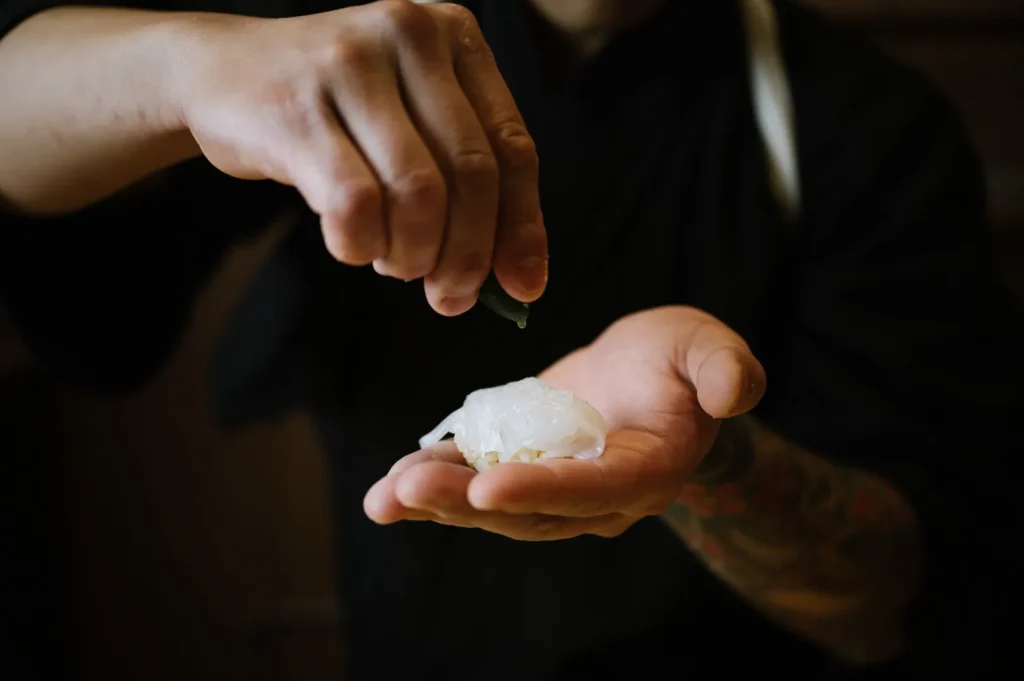 A person in a black garment is delicately squeezing lime juice over fresh sushi held in their tattooed hand, conveying a calm, precise atmosphere.