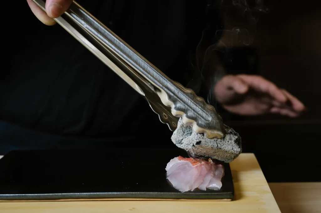 Chef Masa holds hot charcoal with tongs, roasting raw fish on a black plate. Steam rises, creating an atmosphere of culinary precision and innovation.