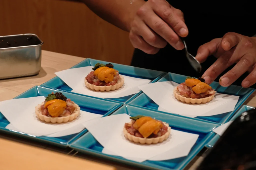 Chef preparing gourmet tartlets with sea urchin on blue plates, conveying elegance and precision. The scene has a focus on fine dining presentation.