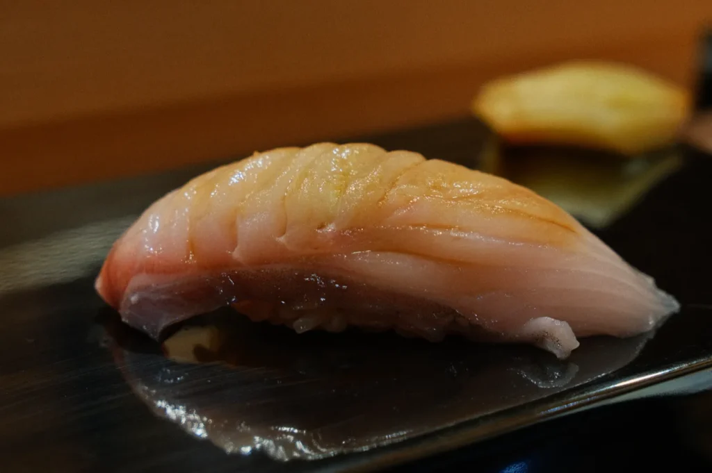 A piece of glistening nigiri sushi with pale pink fish on top of white rice sits on a dark reflective plate. The setting is calm and inviting.