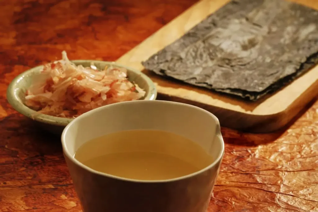 A ceramic bowl of clear broth is in the foreground. Behind it, a plate of bonito flakes and a cutting board with kombu seaweed lie on a textured surface.