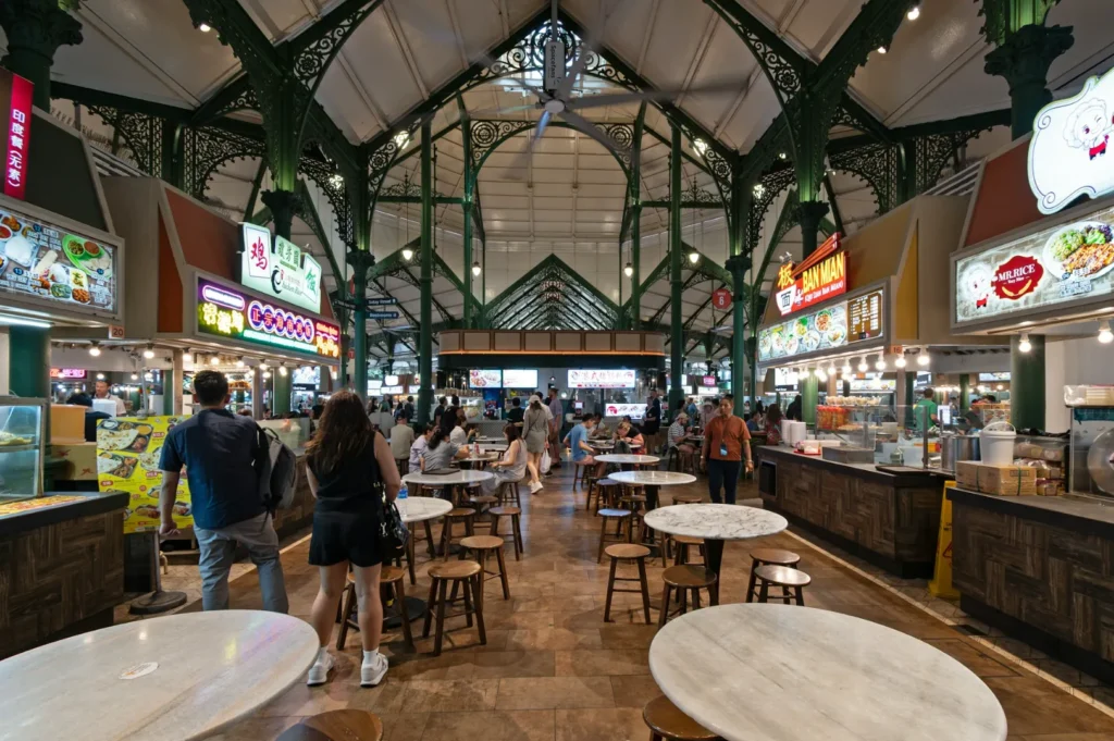 A bustling food court with high ceilings and ornate green iron beams. People browse vibrant food stalls, while others sit at round tables with wooden stools.