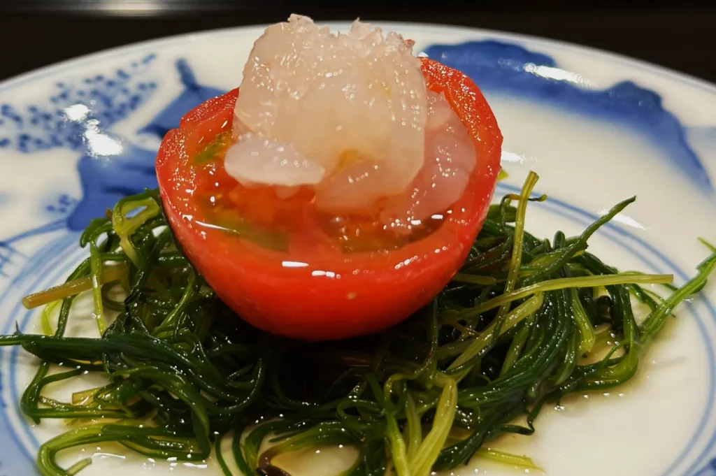 A half tomato filled with translucent jelly sits on a bed of green seaweed on a white plate with a blue pattern, creating a fresh, elegant presentation.