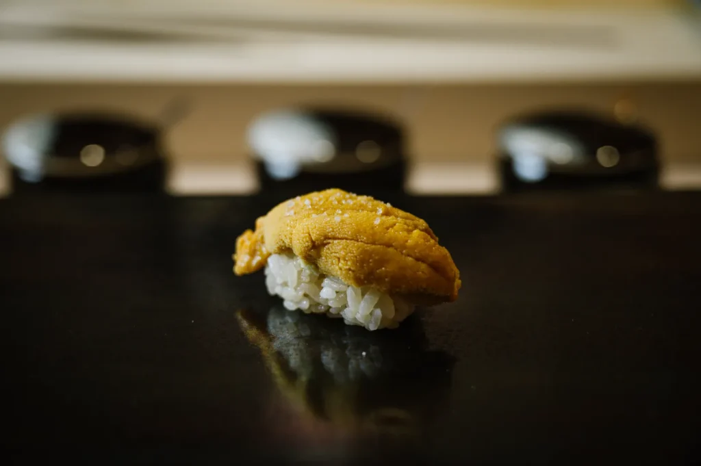 A piece of uni sushi sits elegantly on a dark surface, with glossy rice and bright yellow sea urchin. Blurred background adds a refined ambiance.