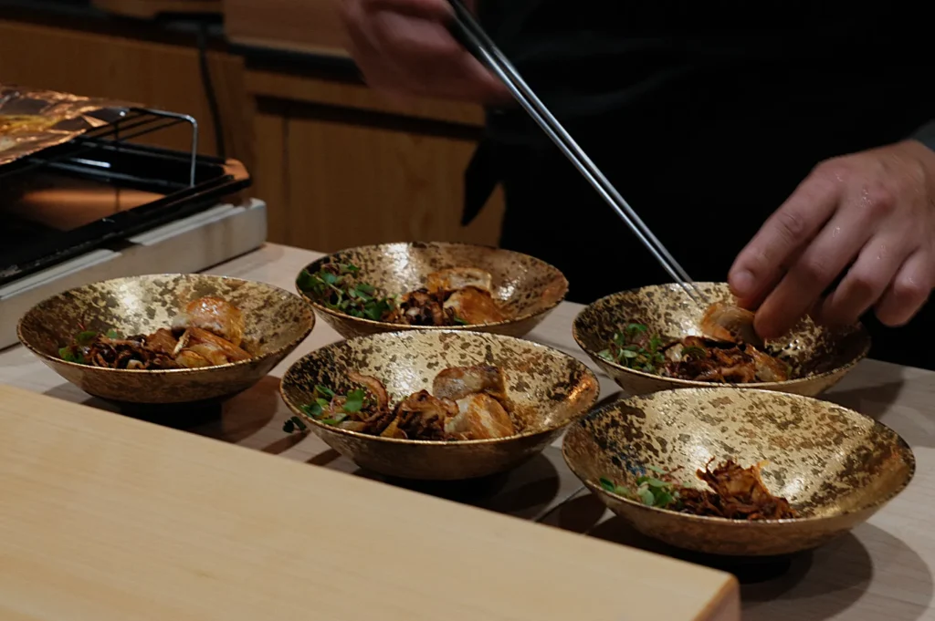 Chef plating five elegant gold bowls with gourmet food, garnishing with greens. The setting is refined, evoking a sense of culinary artistry.