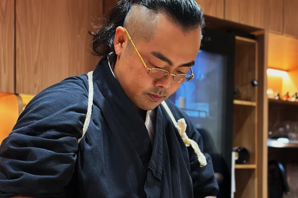 Chef Masa with glasses, wearing a dark traditional robe, is focused on an activity. The background features wooden shelves and soft lighting. Calm and attentive mood.