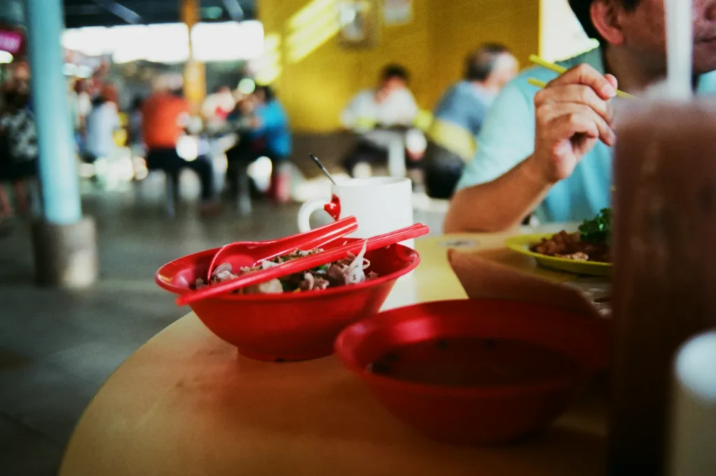 Blurred food court scene with red bowls, noodles, and chopsticks on a table. A person eats nearby, creating a casual, social atmosphere.