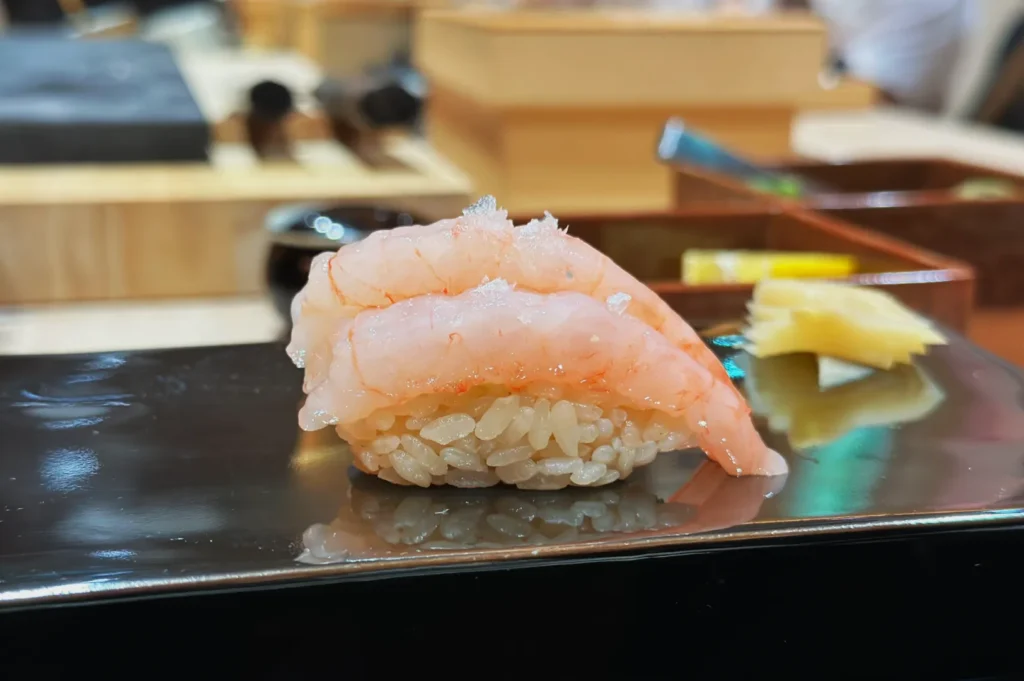 Close-up of a delicate shrimp nigiri sushi on a glossy black plate, garnished with a sprinkle of salt, with ginger slices in the background.