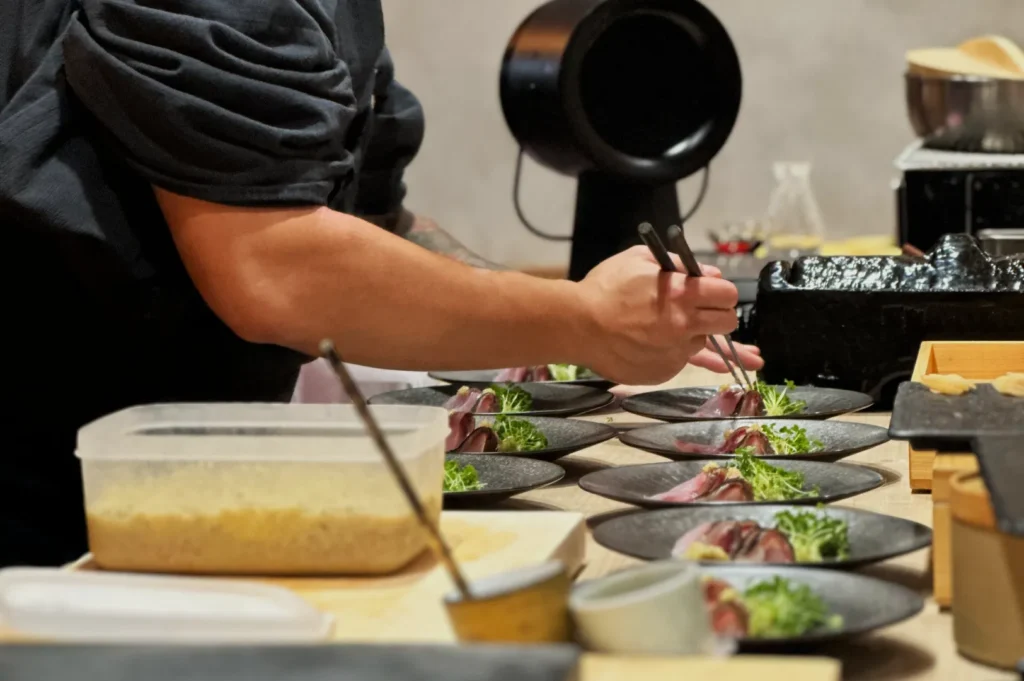 Chef arranging food artfully on multiple black plates, each garnished with greens, on a wooden counter. The scene conveys a focused, creative atmosphere.