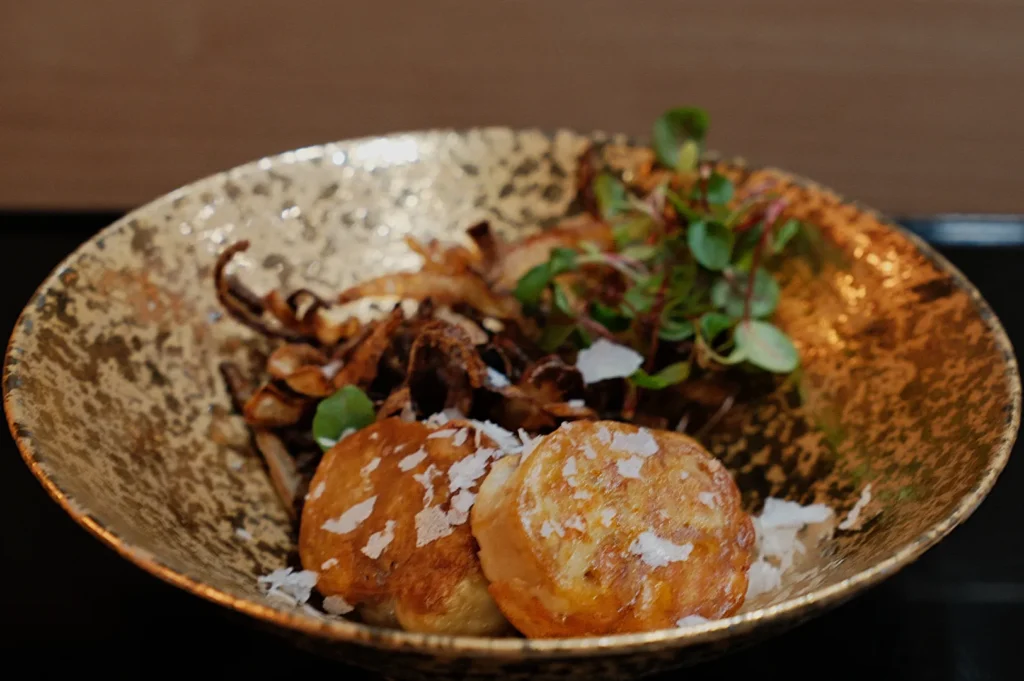 A rustic golden bowl holds two flaky pastries topped with cheese flakes, beside a garnish of microgreens and crispy fried onions. Warm, gourmet presentation.