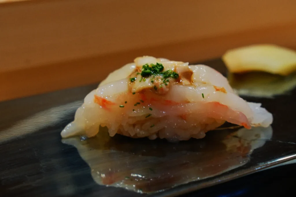 Close-up of a sushi piece with translucent white fish, garnished with green herbs on a sleek, dark plate. The setting is elegant and minimalistic.