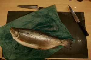 A whole fish on a green sheet of plastic, with knives nearby on a black cutting board.