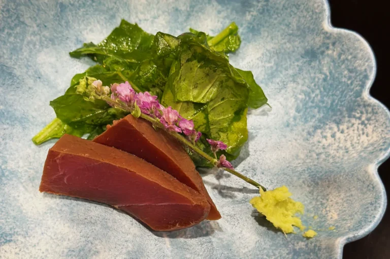 Sliced tuna sashimi garnished with pink flowers and a green salad, served on a textured blue plate. A wasabi dollop adds a spicy touch.