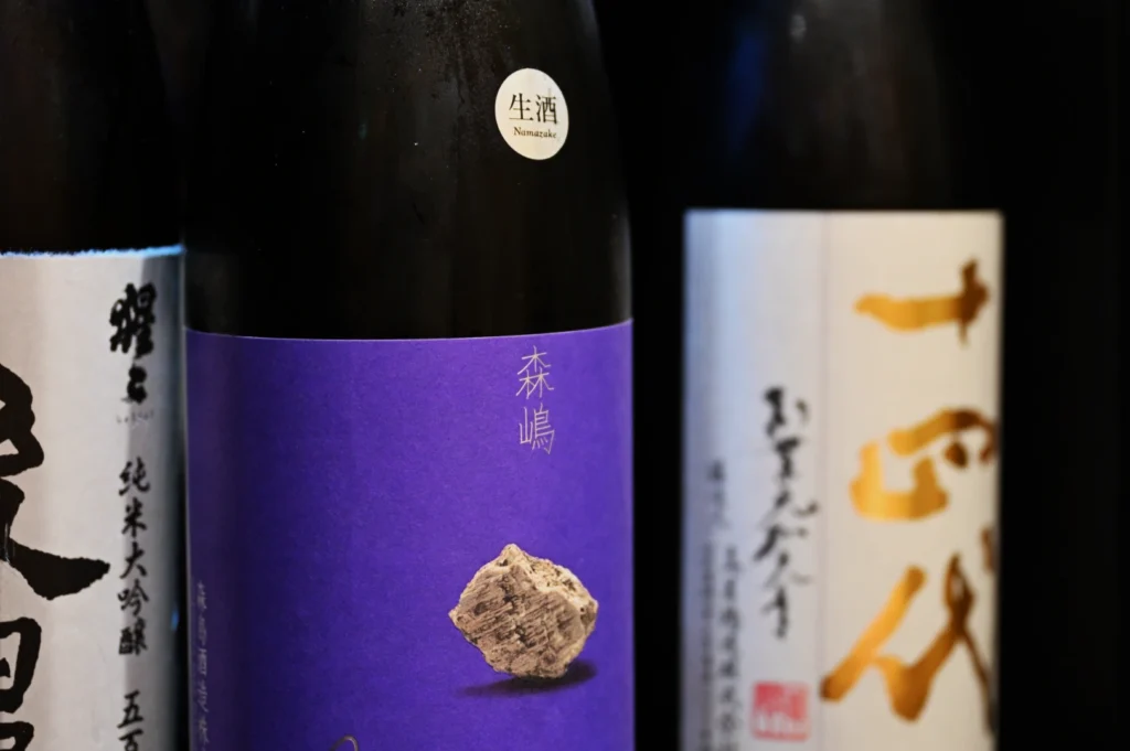 Three bottles of sake, each with distinct labels: one is white with black text, another has a purple label with a stone graphic, and the third features stylized characters on a light background.