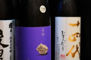 Three bottles of sake, each with distinct labels: one is white with black text, another has a purple label with a stone graphic, and the third features stylized characters on a light background.