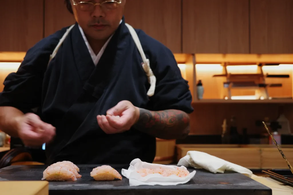 Chef Masa in a traditional outfit preparing sushi on a wooden counter, with several sushi pieces displayed.