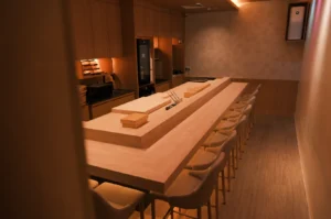 Warm, minimalist sushi bar with light wood interiors. Center island has knives and trays, surrounded by neatly arranged beige chairs, creating a serene ambiance.