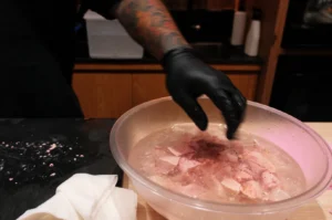 A tattooed hand in black gloves handles raw meat in a clear bowl filled with water. A white cloth lies nearby on a black surface, creating a focused kitchen scene.