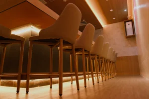 A row of empty, modern bar stools line a warmly lit bar counter with wood accents. The setting conveys a cozy, inviting, and elegant atmosphere.