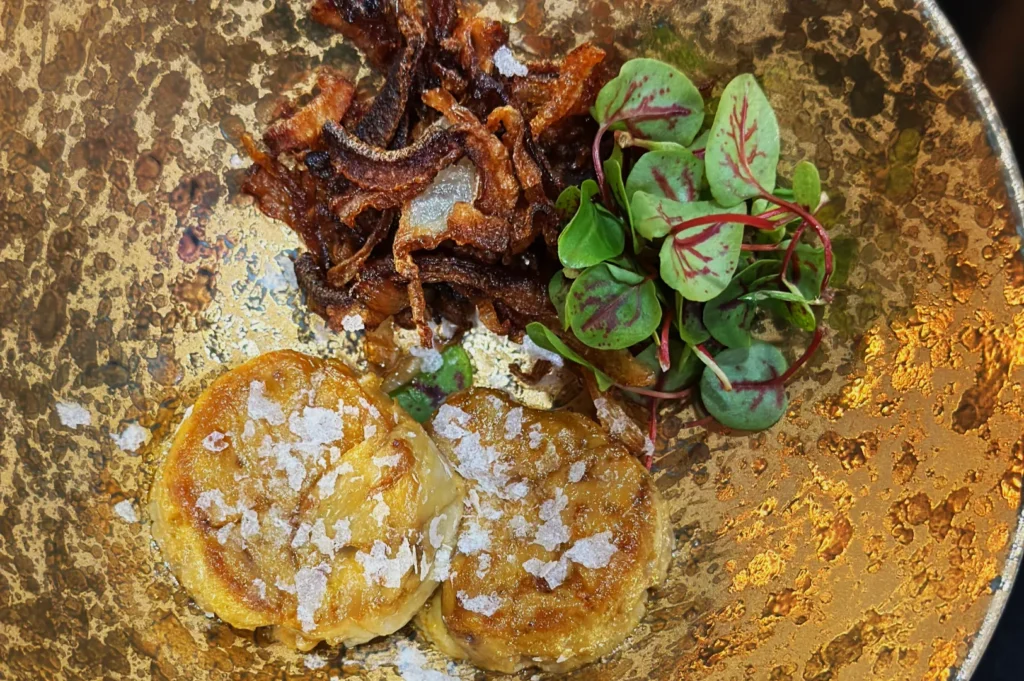 A rustic plate features two caramelized scallops sprinkled with salt, crispy fried onions, and fresh green sprouts, creating a contrast of textures.