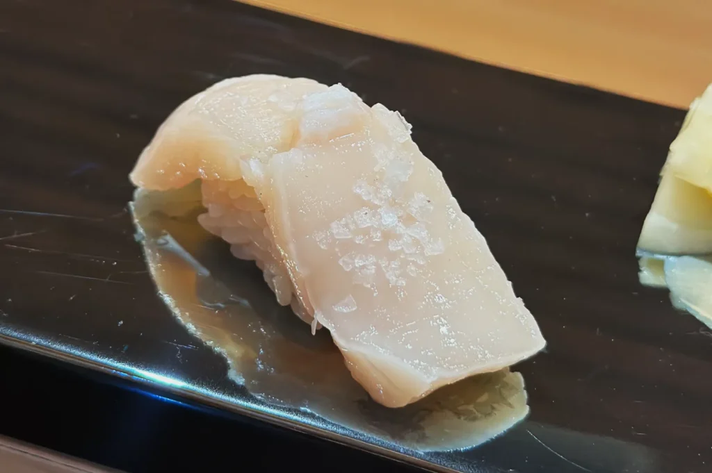 A piece of sushi featuring a slice of white fish atop rice, presented on a black plate.