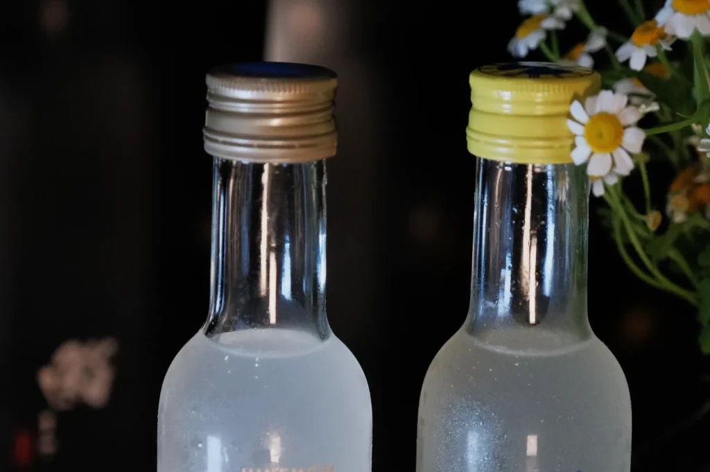 Two glass bottles with metal and plastic caps, partially filled with a clear liquid, next to a small bouquet of white flowers.