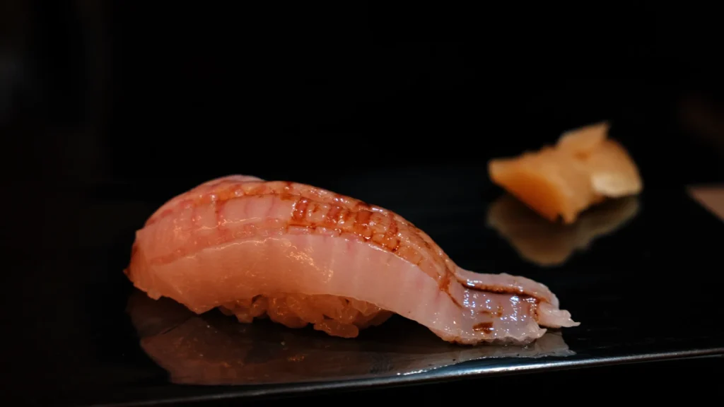 A piece of sushi with a lightly grilled fish on top, served on a black plate with a piece of ginger on the side.