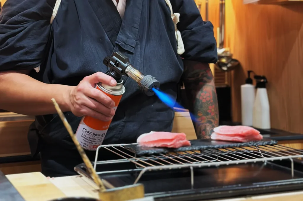 Chef Masa using a culinary torch to sear meat on a grill, with two pieces of meat positioned on a rack.